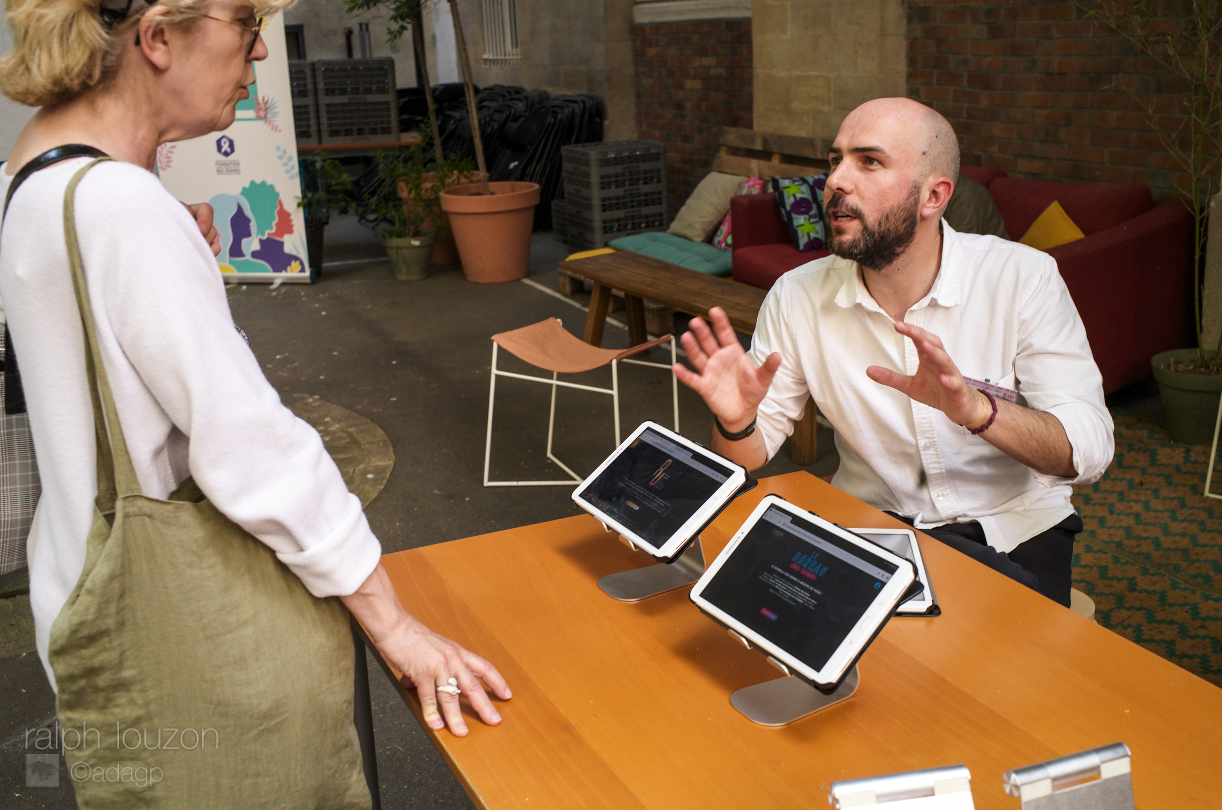 Jean Figarol est assis derrière une table sur laquelle est posée 2 tablettes présentant des serious games. Il explique l'un d'eux à une invitée debout devant.